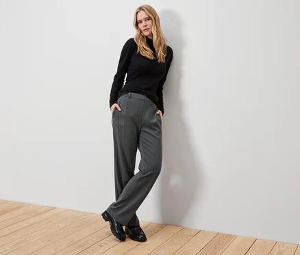 Blonde Frau posiert in schwarzem Rollkragenpullover, grauer Karohose und schwarzen Schuhen.