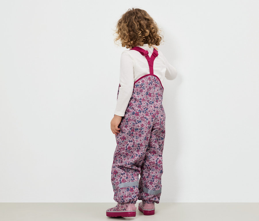 Mädchen in blumiger Latzhose und rosa Stiefeln steht mit dem Rücken zum Betrachter.