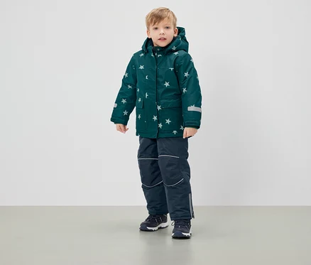 Junge steht in grüner Winterjacke mit Sternen und dunkelblauer Hose.