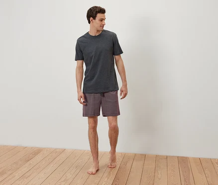 Mann in grauem T-Shirt und gemusterter Shorts steht barfuß auf Holzboden vor einer weißen Wand.