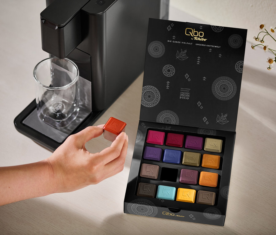 Eine Hand hält eine rote Kaffeekapsel neben einer schwarzen Qbo ICONIC Dark Chrome Kaffeemaschine mit einem Glasbecher und einer offenen Qbo Collection - 16 Kapseln Box mit bunten Kapseln.