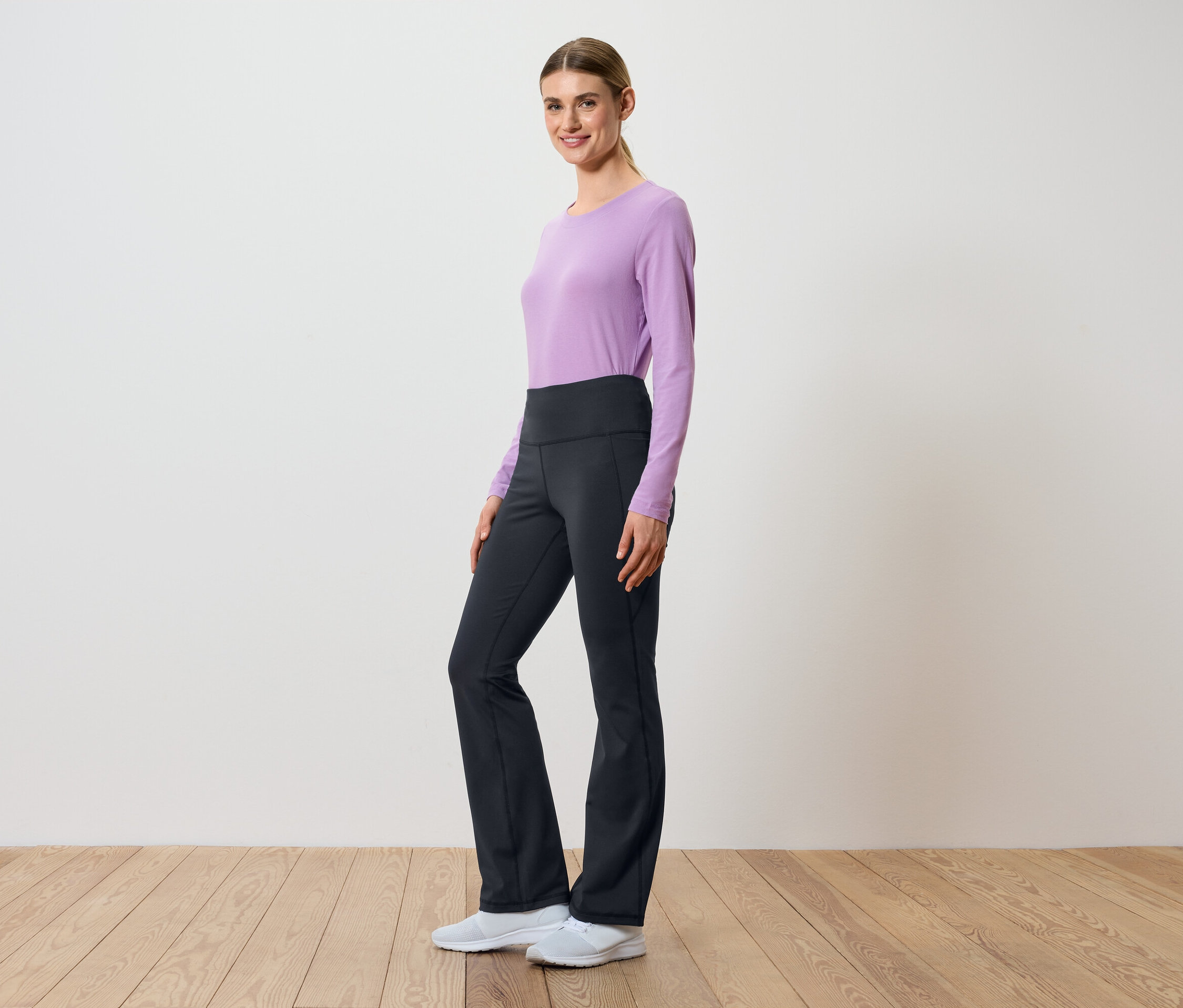 Eine Frau posiert seitlich in einem lila, langärmeligen Oberteil und einer schwarzen Hose mit hohem Bund.