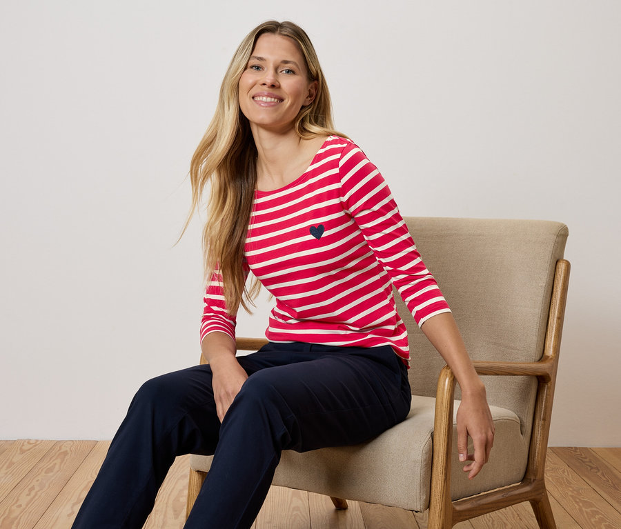 Eine Frau sitzt auf einem Stuhl und trägt ein rot-weiß gestreiftes T-Shirt und dunkelblaue Hosen.