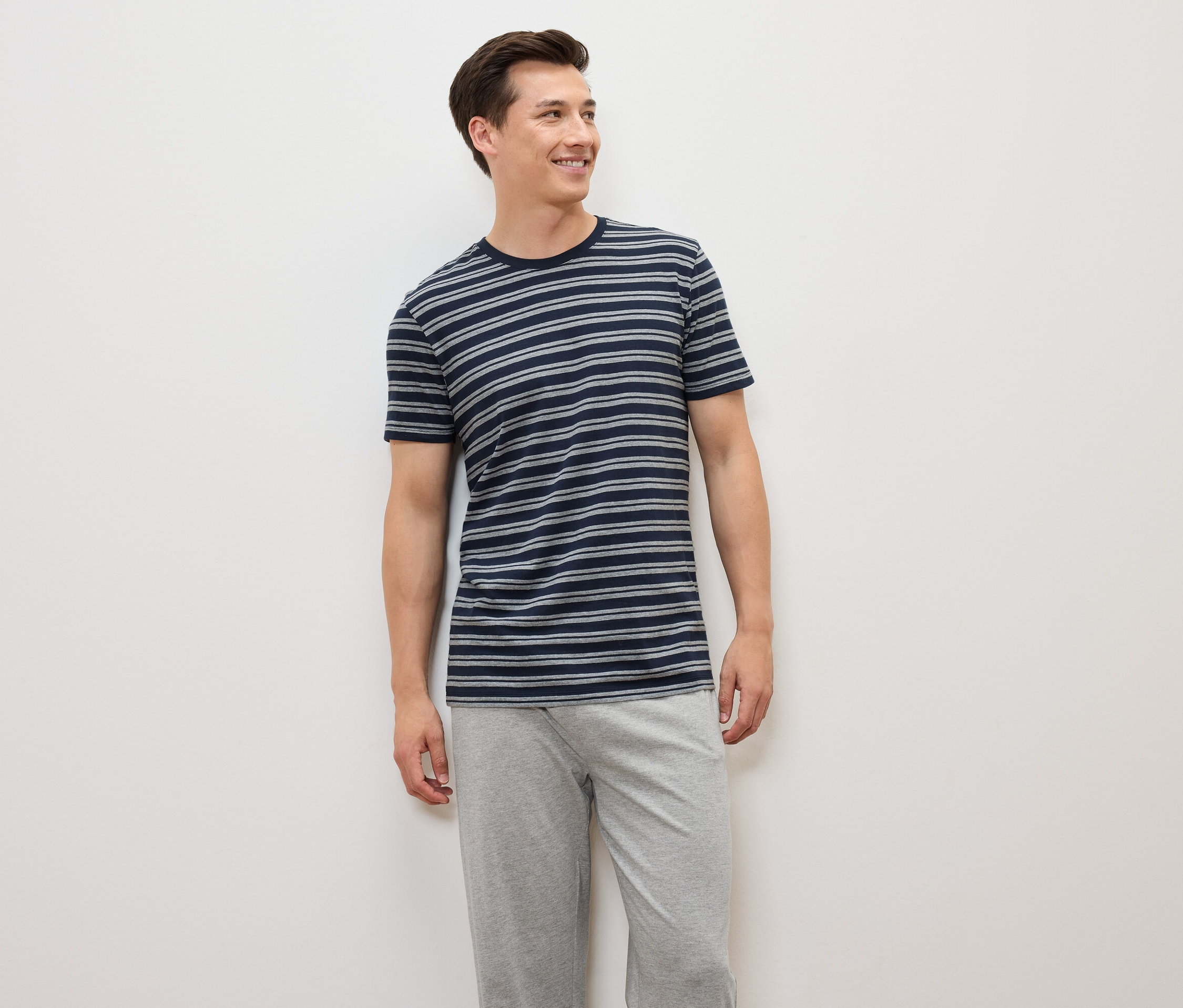 Ein Mann steht und trägt ein blau-grau gestreiftes T-Shirt und graue Hosen.