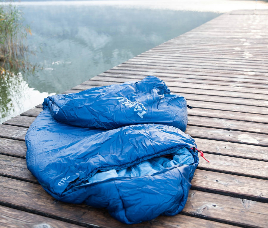Blauer Tambu Mumienschlafsack Talu 1500 liegt auf einem Holzsteg am See.