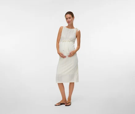 Schwangere Frau trägt ein ärmelloses, weißes Kleid mit einem Band um die Taille und Flip-Flops.