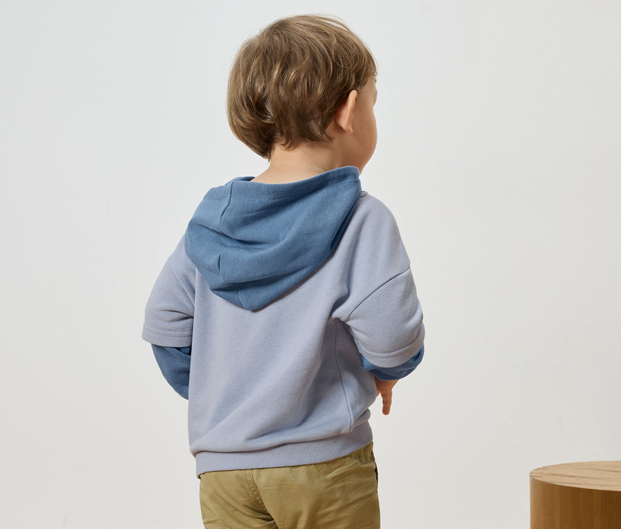 Rückansicht eines Kindes mit blonden Haaren, das einen blau-grauen Kapuzenpullover und eine Hose trägt.