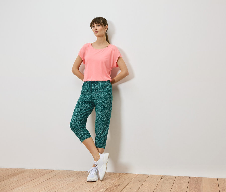 Frau lehnt an einer Wand, trägt ein korallfarbenes T-Shirt, grüne Caprihosen mit Blattmuster und weiße Turnschuhe.