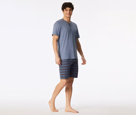 Ein lächelnder Mann trägt ein blaues SCHIESSER Shorty-Set mit Kurzarm-Henley-Oberteil und gestreiften Shorts, barfuß stehend.