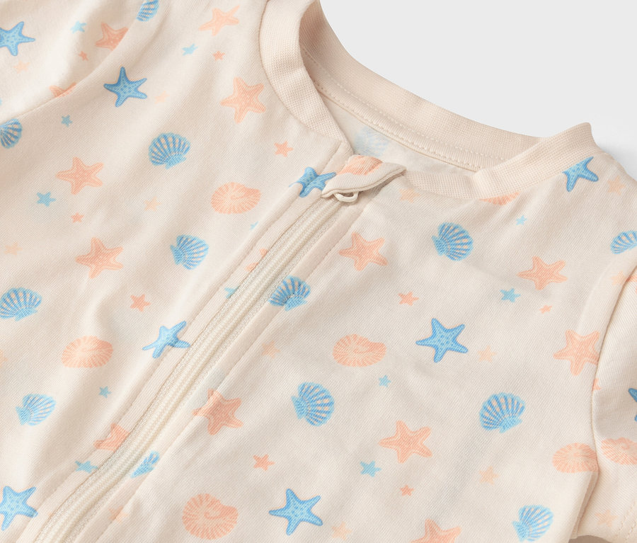 Nahaufnahme eines cremefarbenen Baby-Schlafanzugs mit Reissverschluss und einem Allover-Muster aus blauen Muscheln und Seesternen in Pfirsichfarbe.