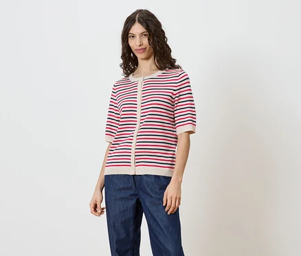 Frau mit braunem, gewelltem Haar trägt eine gestreifte, kurzärmelige Strickjacke mit Knöpfen und Jeans vor einem weißen Hintergrund.