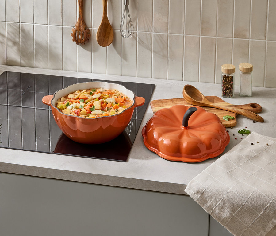 Orangefarbener Kürbis-Topf mit Deckel steht auf einem Herd neben einem Holzlöffel und Gewürzen.