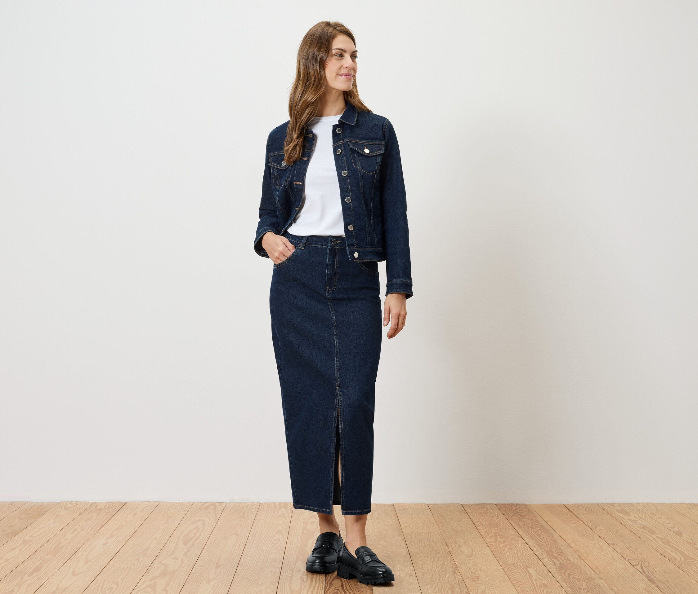 Eine Frau steht und trägt eine Jeansjacke und einen Jeansrock mit Schlitz.
