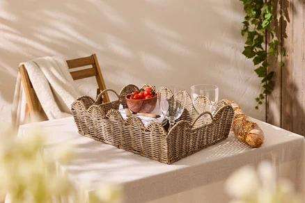 Auf einem Tisch mit Tischdecke steht ein geflochtenes Tablett mit Tomaten, Gläsern und Baguette.
