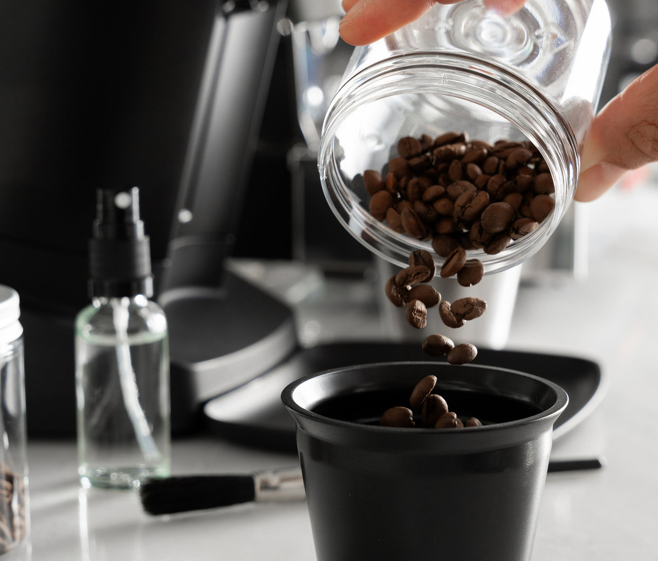 Eine Hand schüttet Kaffeebohnen aus einem Glasbehälter in eine schwarze Tasse.