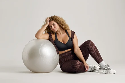 Frau mit lockigem Haar sitzt neben einem silbernen Gymnastikball, trägt einen schwarzen Sport-BH, Leggings mit Zebramuster und Turnschuhe.