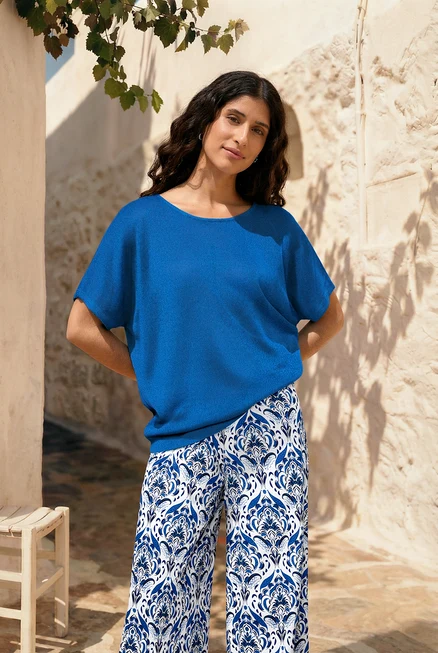 Frau mit Creolen Pavé, blauem, strukturiertem Shirt und Palazzohose mit Alloverprint vor einer Wand.
