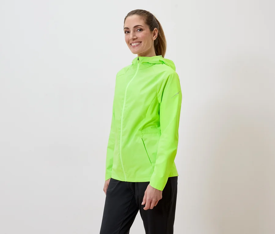Frau mit Pferdeschwanz trägt eine neongrüne Regenjacke und schwarze Hose vor einem weißen Hintergrund.