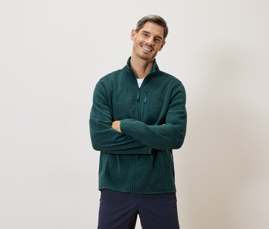 Mann mit verschränkten Armen, trägt eine grüne Fleecejacke mit Reißverschluss und marineblaue Shorts vor einem hellen Hintergrund.