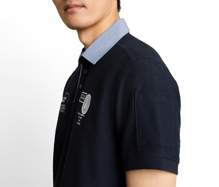 Nahaufnahme eines Mannes in einem dunkelblauen Poloshirt mit hellblauem Kragen, weißer Stickerei auf der Brust und einer kleinen Taschendetail am Ärmel.