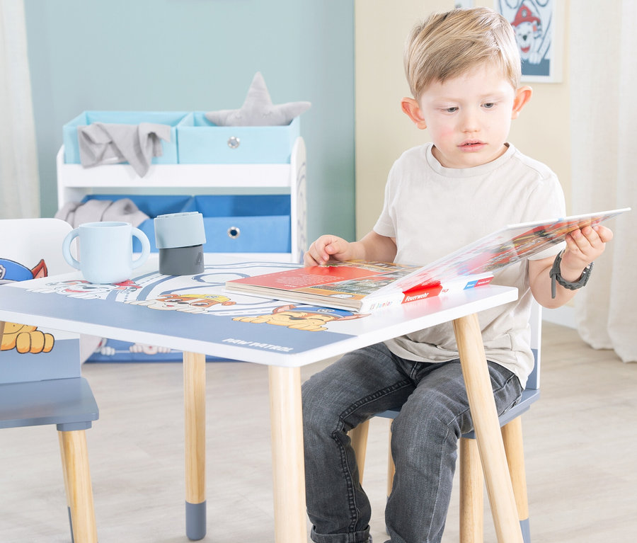 Ein blonder Junge sitzt an einem Kindertisch mit Paw Patrol-Motiv und liest ein Buch.