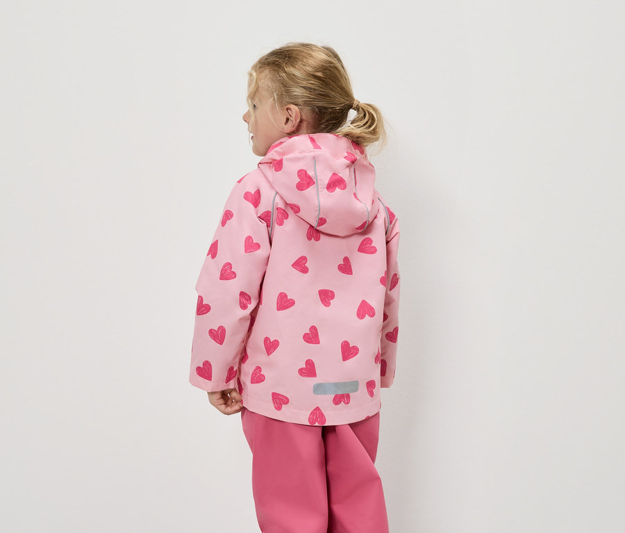 Rückansicht eines kleinen Mädchens mit blonden Haaren, das eine rosafarbene Regenjacke mit Herzmuster und eine passende Hose trägt.