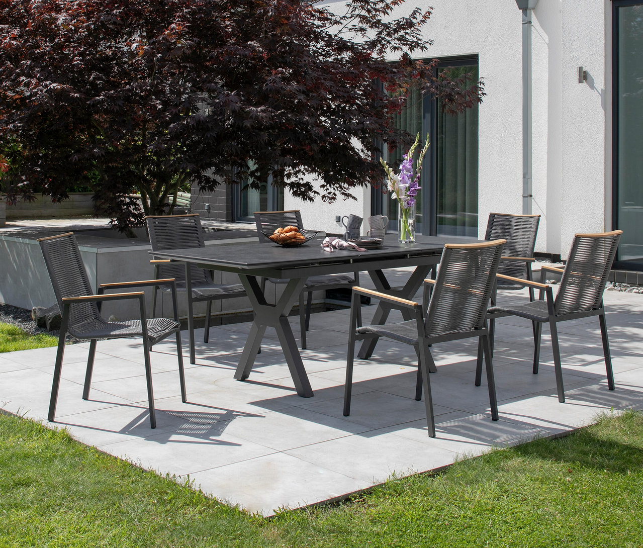 Grauer Gartentisch mit sechs Stühlen auf einer Terrasse mit einem Baum.