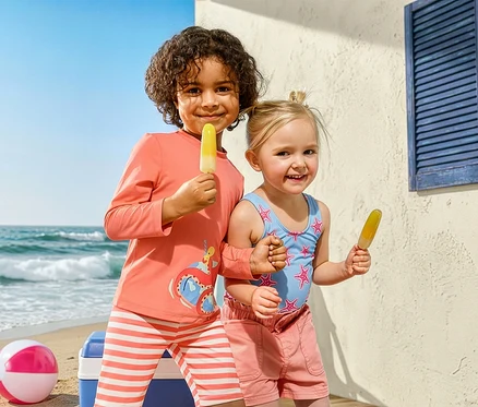 Zwei Kinder mit Eis am Strand. Das Mädchen trägt einen Kinder-Badeanzug mit Seesternen und Jeansshorts. Der Junge trägt eine Kinder-Kombi mit UV-Schutz und Krake.