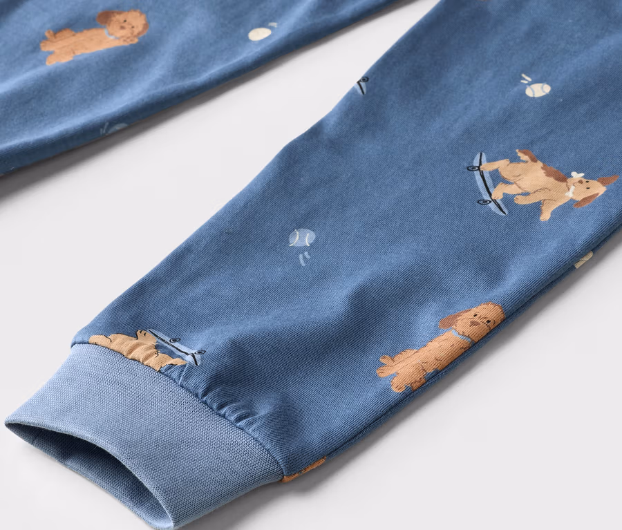 Nahaufnahme eines blauen Stoffes mit einem Muster aus braunen Cartoon-Hunden und weißen Tennisbällen. Ein Hund ist auf einem Skateboard. Der Stoff hat ein geripptes Bündchen.