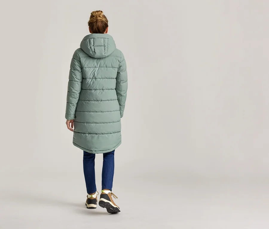 Rückansicht einer Frau mit Duttfrisur, die einen langen, mintgrünen Steppmantel mit Kapuze, Jeans und Wanderschuhe trägt.
