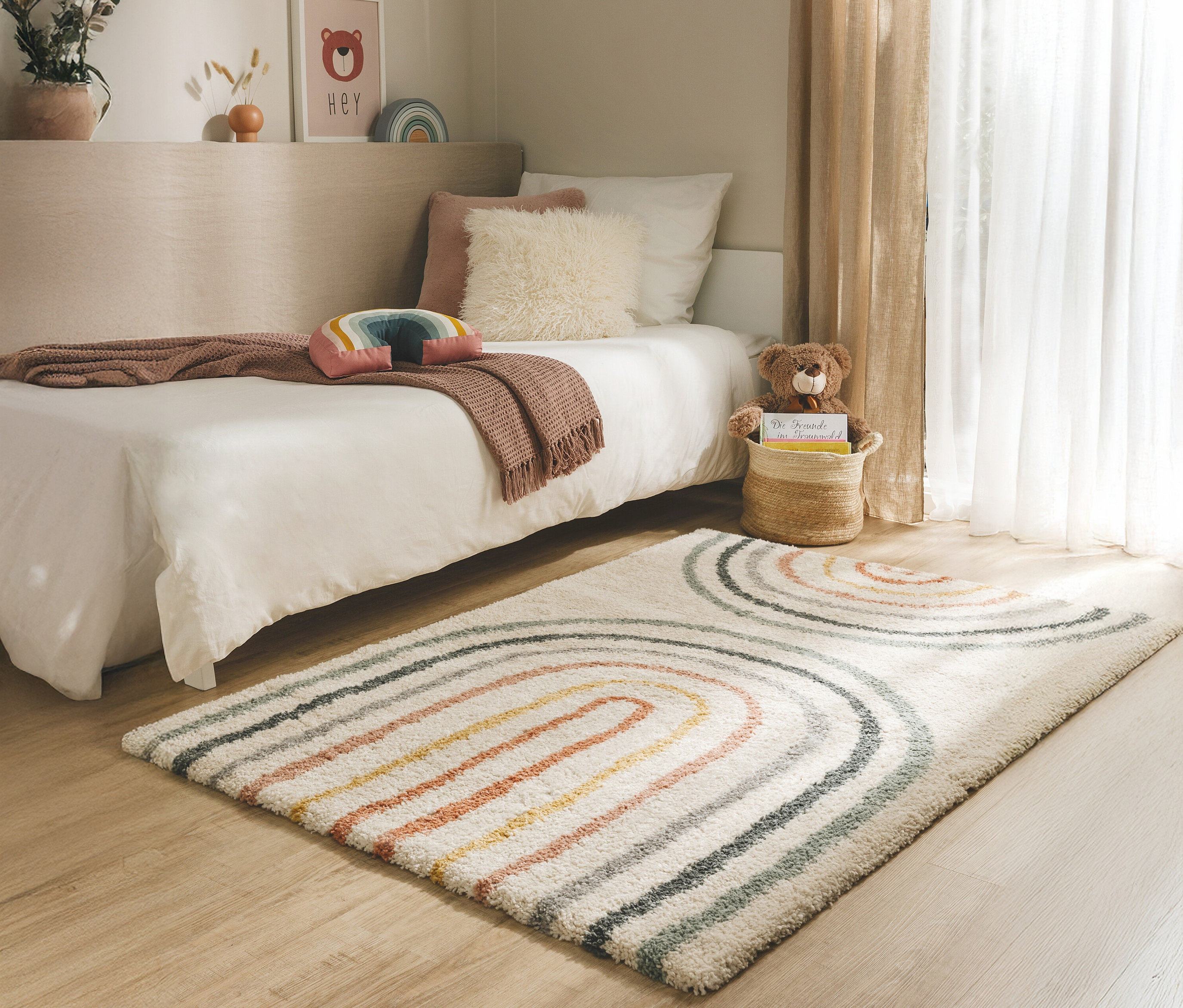 Ein Kinderzimmer mit einem Bett, einem Teppich mit Regenbogenmuster und einem Teddybären in einem Korb.