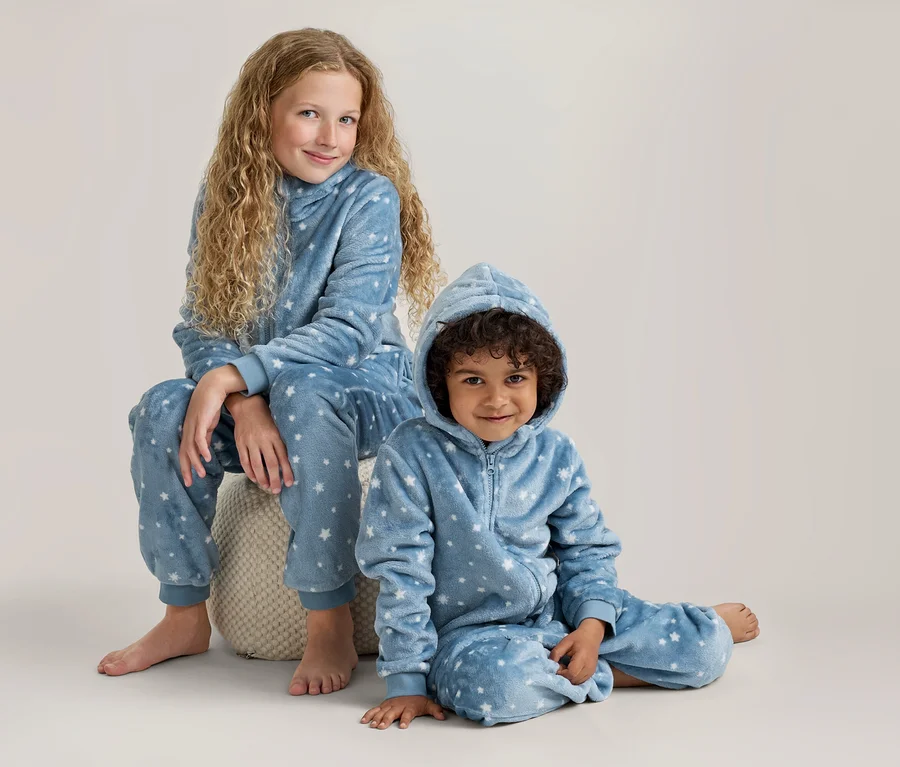 Zwei Kinder, ein älteres Mädchen und ein jüngerer Junge, tragen passende blaue Fleece-Schlafanzüge mit Kapuze und weißen Sternen. Das Mädchen sitzt auf einem hellen Pouf, der Junge sitzt am Boden, beide lächeln.