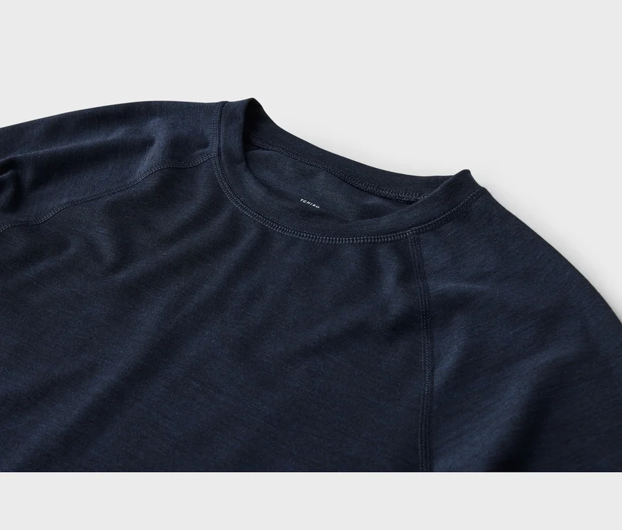 Nahaufnahme eines dunkelblauen Langarmshirts mit Rundhalsausschnitt und Raglanärmeln. Der Stoff hat eine leicht melierte Textur, sichtbare Nähte verlaufen entlang des Ausschnitts und der Ärmelnähte. Ein weißes „TCHIBO“-Etikett ist im Kragen sichtbar.