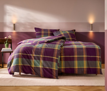 Ein Bett mit lila, grün, gelb und braun karierter Bettwäsche, bestehend aus einem Bettbezug und zwei Kissen. Links daneben steht ein Holzstuhl mit einem Stapel Bücher und einer grünen Vase mit Blumen. Die Wand hinter dem Bett ist zweifarbig gestrichen, unten dunkelviolett und oben hellrosa-violett, mit zwei Wandleuchten. Ein heller Teppich liegt auf dem Holzboden.