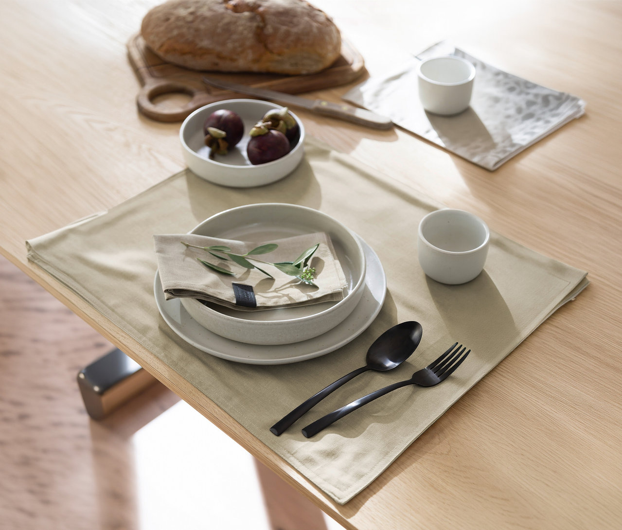 Ein Esstisch ist gedeckt mit einem sandfarbenen SCHÖNER WOHNEN-Kollektion Tischset, weißen Tellern, einer Leinenserviette mit grünem Zweig und schwarzem Besteck. Im Hintergrund sind ein Brotlaib auf einem Holzbrett, eine Schale Mangostanen und eine weitere weiße Tasse zu sehen.