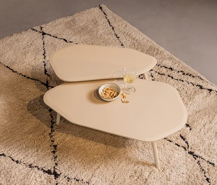 Zwei unregelmäßig geformte, hellbeige Couchtische stehen auf einem zotteligen, hellen Teppich mit schwarzen Mustern. Auf dem unteren Tisch sind eine Schale Erdnüsse, ein Glas Weißwein und Erdnussschalen.