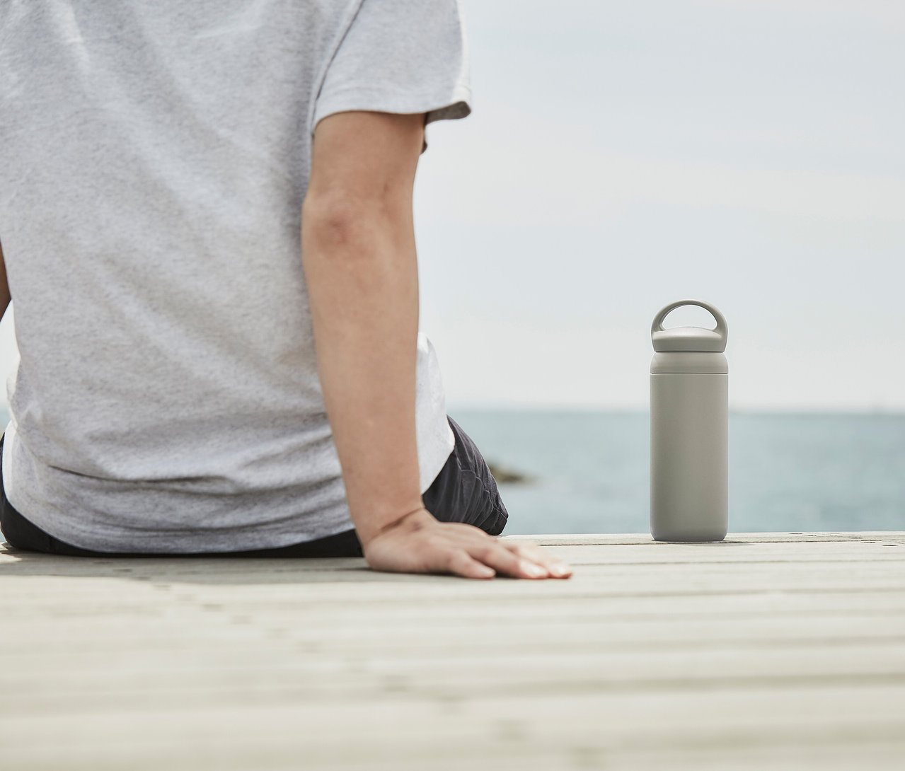 Person sitzt auf Holzsteg, daneben steht ein beiger KINTO Tumbler »Day Off« vor dem Meer.