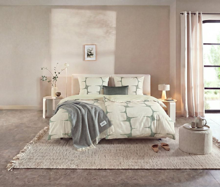 Ein Schlafzimmer mit einem Bett, das mit der SCHÖNER WOHNEN – Kollektion Bettwäsche in Hellgrün und Weiß mit geometrischem Muster bezogen ist. Ein graues Plaid liegt am Fußende, daneben ein Hocker mit Teekanne und Tassen.