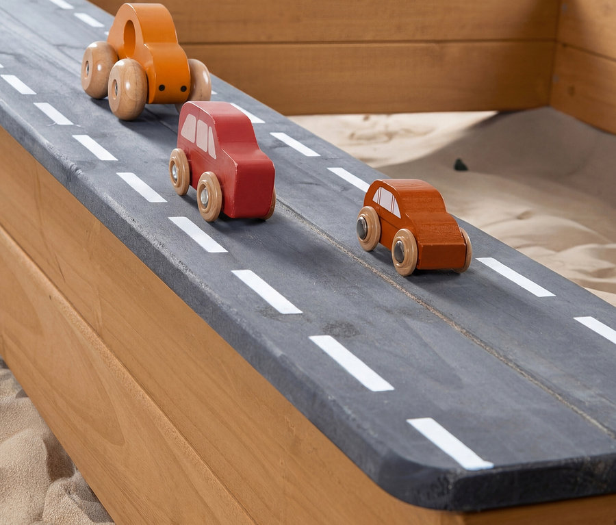 Drei hölzerne Spielzeugautos (orange, rot, braun) auf einer dunkelgrauen Holzstraße mit weißen gestrichelten Linien, eingebaut in einen Holzsandkasten.