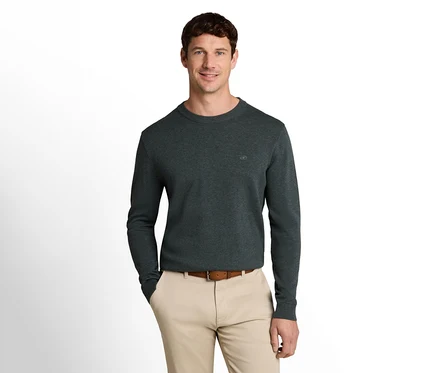 Lächelnder Mann in dunkelgrünem Langarmpullover, hellbeiger Hose und braunem Gürtel, eine Hand in der Hosentasche.