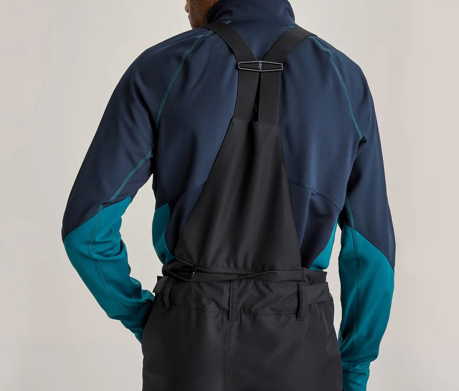 Rückenansicht einer Person in einem dunkelblauen und türkisfarbenen Langarmshirt und schwarzer Latzhose mit Hosenträgern. Ein Reißverschluss ist am Rücken der Hose sichtbar.