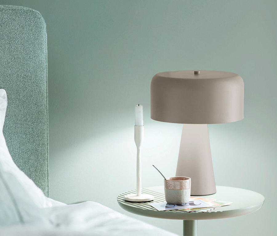 Runder, hellgrüner Nachttisch neben einem Bett mit grünem Kopfteil. Darauf eine SCHÖNER WOHNEN-Kollektion Tischleuchte »Pento« in Sandbrise, ein weißer Kerzenständer und eine zweifarbige Tasse.