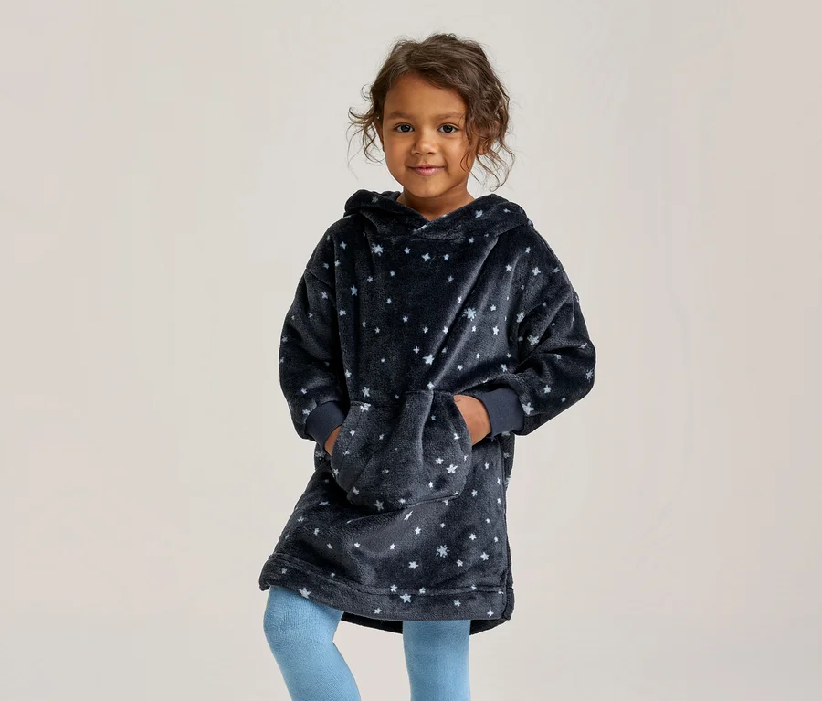 Ein junges Mädchen lächelt, trägt einen dunkelblauen, übergroßen Kapuzenpullover mit weißen Sternen und hellblauen Leggings, die Hände in der Vordertasche.