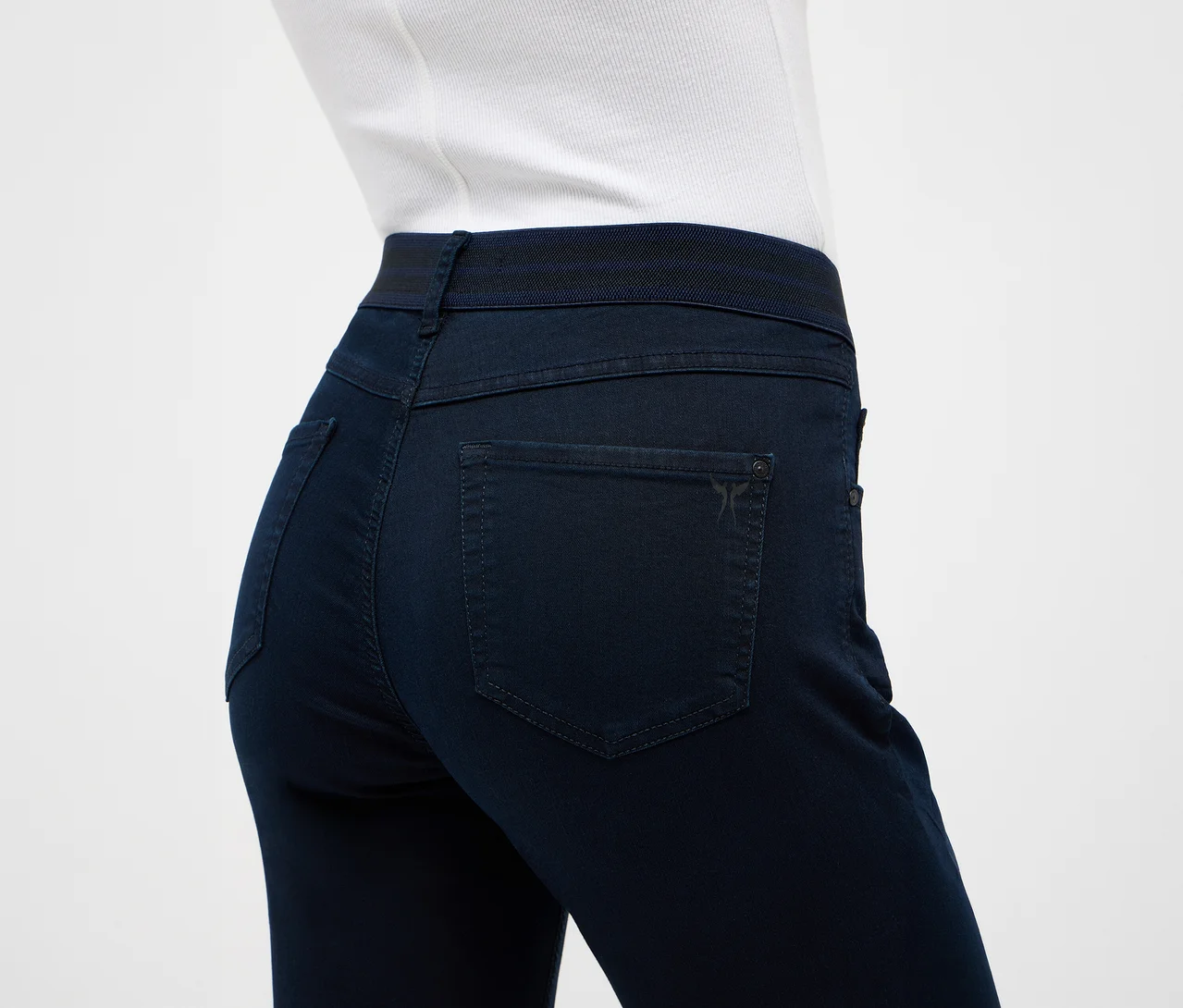 Rücken einer Person in dunkelblauen Jeans mit elastischem Bund und weißem Oberteil. Die Jeans hat Gesäßtaschen, eine mit kleinem dunklen Logo.