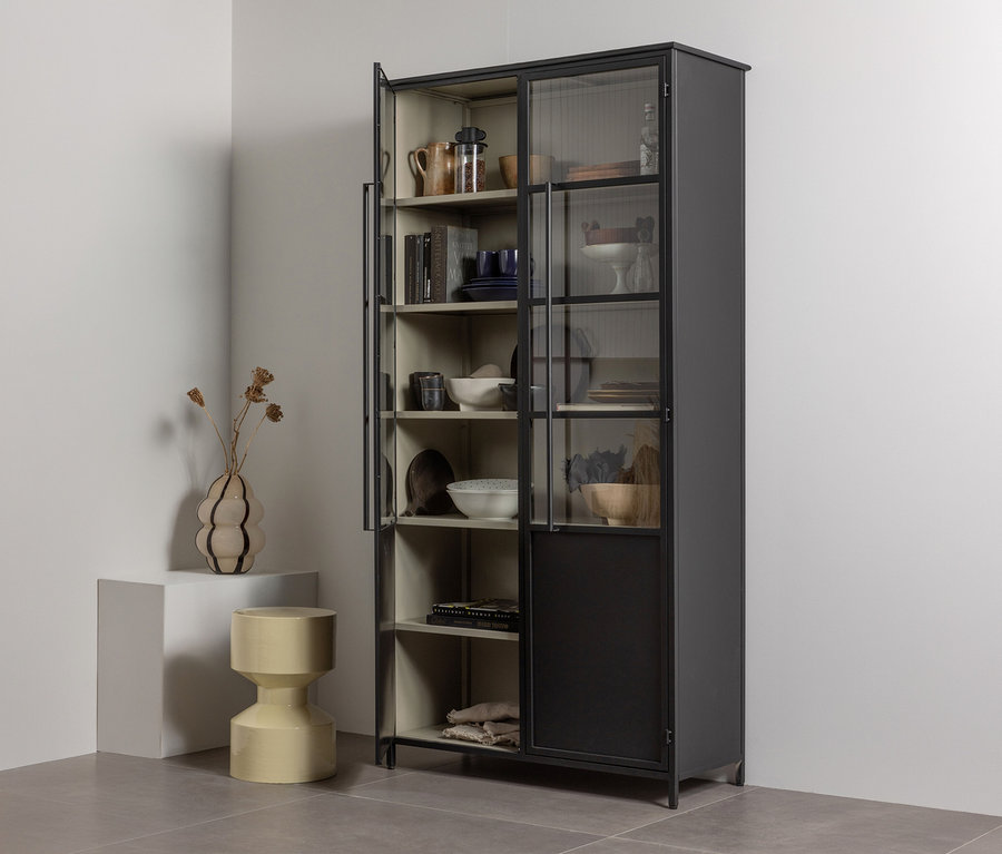 Dunkelgrauer Vitrinenschrank mit Glastüren, gefüllt mit Geschirr und Dekoartikeln, steht in einer Raumecke. Links davon ein weißer Sockel mit einer Vase mit Trockenblumen und ein gelber Hocker.