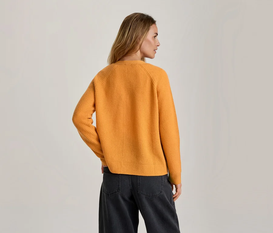 Frau von hinten, leicht nach rechts gedreht, trägt einen orangefarbenen Rippstrickpullover und eine dunkelgraue weite Jeans.