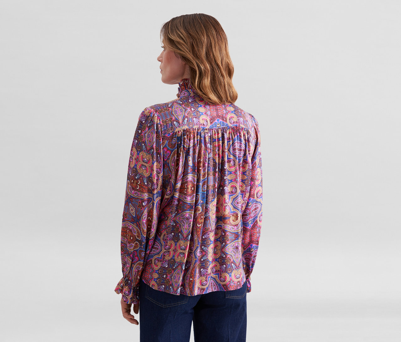 Frau von hinten, trägt eine langärmelige Bluse mit buntem Paisleymuster und Rüschenkragen sowie dunkle Jeans.