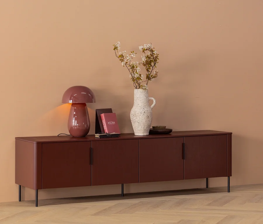 Ein dunkelbraunes Sideboard mit vier Türen steht vor einer beigen Wand. Darauf stehen eine leuchtende pilzförmige Lampe, zwei Bücher, eine strukturierte cremefarbene Vase mit weißen Blüten und eine kleine Schale.