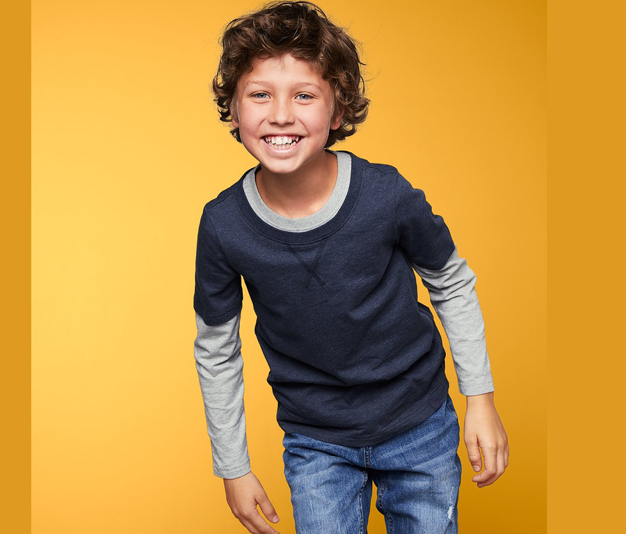 Ein lächelnder Junge trägt ein blaues Langarmshirt und Jeans vor einem gelben Hintergrund.