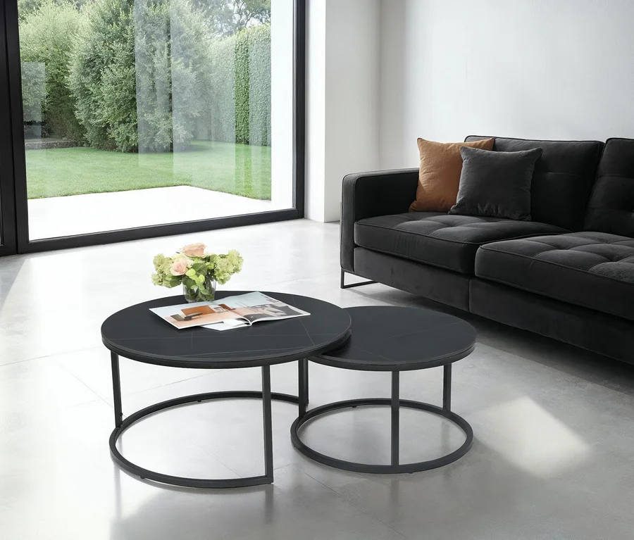 Modernes Wohnzimmer mit schwarzem Sofa und zwei runden, schwarzen Beistelltischen. Auf dem größeren Tisch stehen Blumen und eine Zeitschrift. Ein großes Fenster blickt auf einen grünen Garten.